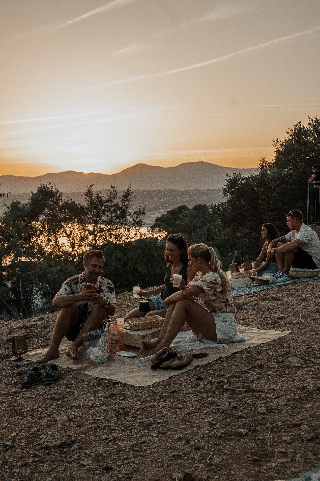 Friends sharing drinks on jute rugs at sunset, hills of the Riviera behind
